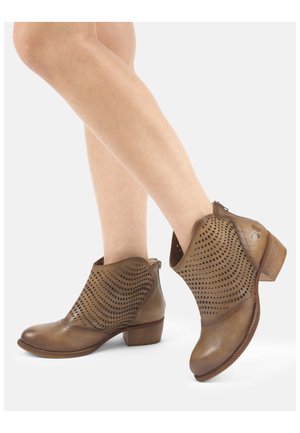 Brązowe skórzane botki do kostki z perforowanymi falistymi wzorami, niskimi drewnianymi obcasami i tylnymi suwakami, noszone na skrzyżowanych nogach.