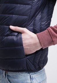 Námořnická puffer vesta s prošívanou texturou, která má boční kapsu. Nosená přes červenou košili s dlouhým rukávem, vystavená na světlém pozadí.