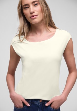 Jonge vrouw met lang blond haar, gekleed in een wit mouwloos topje en donkerblauwe jeans, met handen in de zakken, neutrale achtergrond.