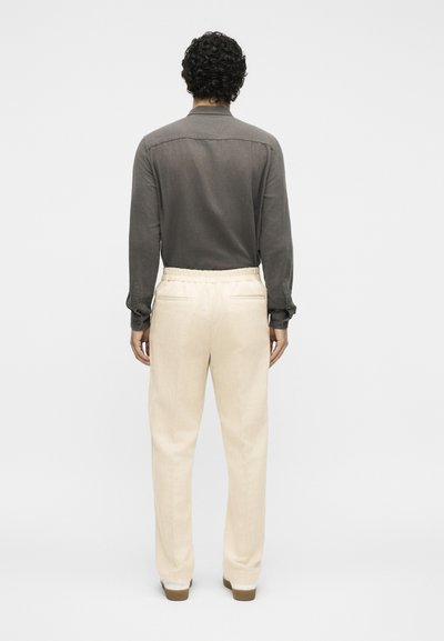 Άνδρας με σγουρά σκούρα μαλλιά, ντυμένος με γκρι μπλούζα μακριάς μανίκιας και μπεζ παντελόνι, στέκεται γυρισμένος προς τα πίσω σε απλό φωτεινό φόντο.