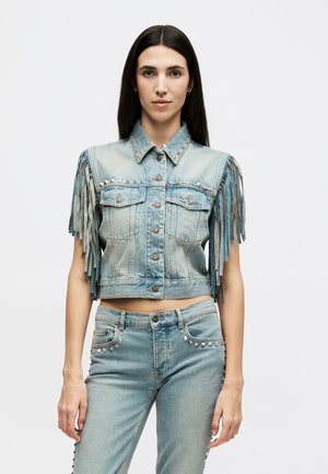 Γυναίκα με ανοιχτό γαλάζιο κοντό τζιν μπουφάν με μανίκια κρόσσια και τζιν παντελόνι με καρφιά που ταιριάζει, στέκεται μπροστά σε λευκό φόντο.