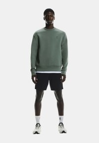 Zielona bluza z okrągłym dekoltem, fakturowana tkanina i detal z logo. Noszona z czarnymi szortami i białymi skarpetkami, w połączeniu z butami sportowymi.