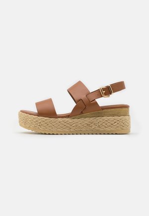 Brun læder platåsandal med to brede remme, en ankelspænde og en flettet jutessål mod en lys baggrund.