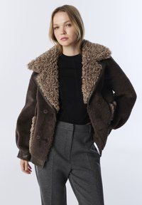 Jeune femme portant une veste en shearling marron foncé avec un large col, un haut noir et un pantalon gris ajusté, se tenant devant un fond clair.