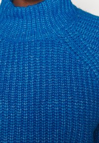 Stickad tröja i livlig blåfärg. Har en hög hals och ribbad textur med vertikala mönster. Mjuk, ullliknande kvalitet.