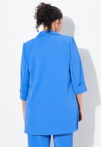 Sininen räätälöity takki, jossa on kaulus, kolmannekselle hihat ja sivuaukot. Valmistettu sileästä kankaasta, jossa on tyylikäs ja rakenteellinen muotoilu.