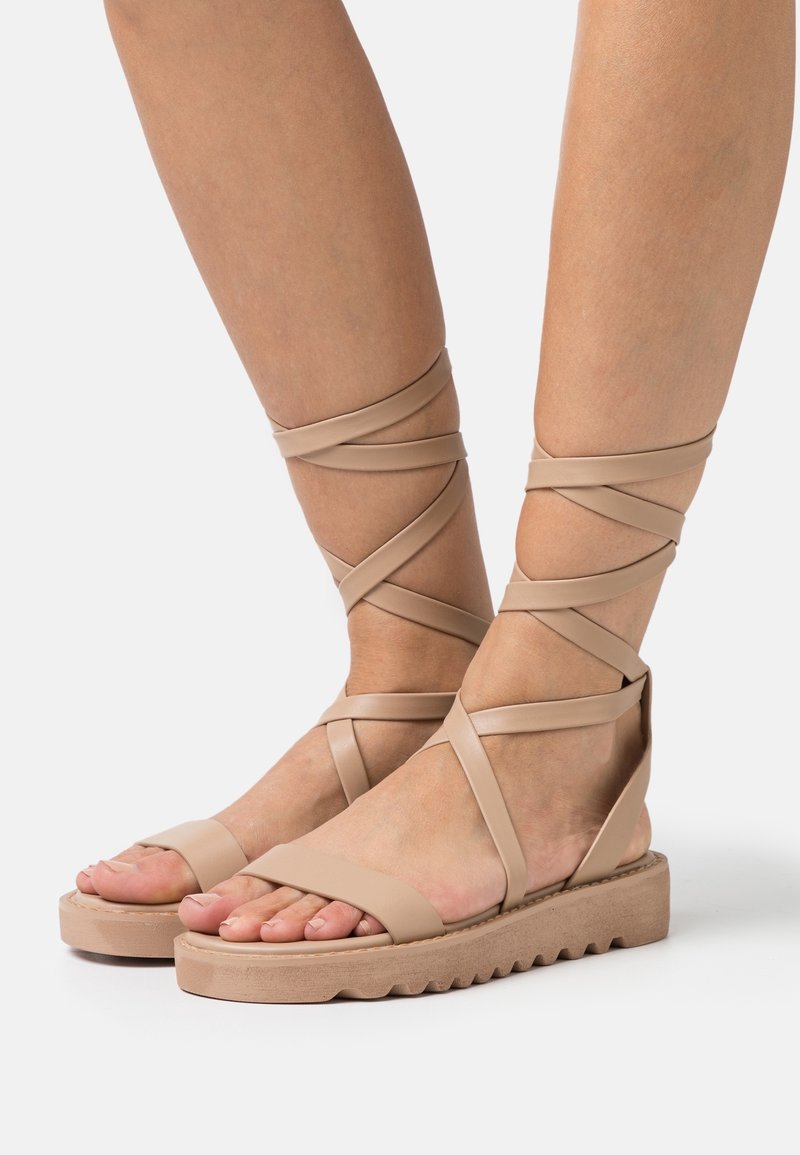Pieds portant des sandales plates beige avec des semelles épaisses et de fines lanières enroulées autour des chevilles sur un fond uni.