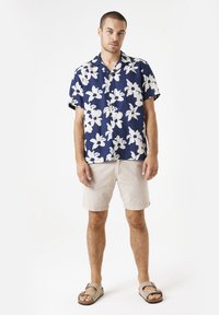 Camisa de manga corta azul marino con patrón floral blanco, combinada con pantalones cortos texturizados de color beige claro y sandalias marrones sin hebillas.