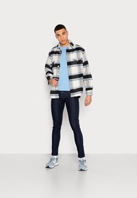 Camisa jaqueta de fleece com vários padrões em preto e branco, sobre uma t-shirt azul. Calças jeans ajustadas azul-escuro e ténis azul-claro.