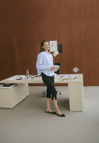 Chemise bleue claire à manches longues, pantalon capri noir avec détail de fente, hauts talons noirs. Bureau minimaliste avec des documents, une bouteille d'eau et une lampe. Mur marron.