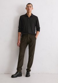 Zwarte polo met lange mouwen, gecombineerd met donkergroene op maat gemaakte broek en zwarte schoenen. Gladde stof met een klassieke kraag en knoopdetails.