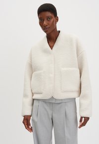 Giacca strutturata color crema con design corto, caratterizzata da due tasche frontali e scollo arrotondato, indossata sopra pantaloni grigio chiaro.