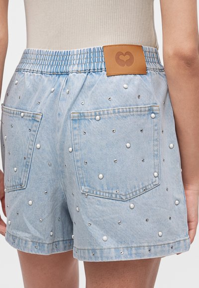 Short en denim bleu clair avec taille élastique, poches arrière et petits clous blancs et argentés éparpillés, porté par une personne en haut beige.