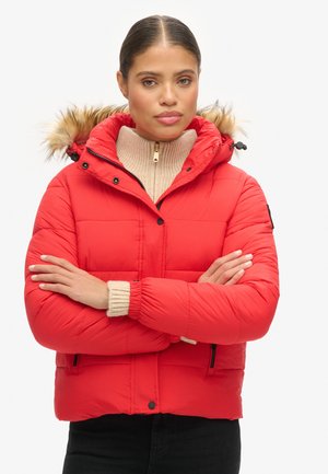 Red puffer jacket with fur-lined hood, quilted design, elastic cuffs, and black zipper pockets. Worn over a beige high-neck sweater.