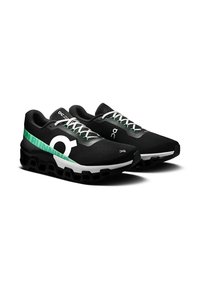 Črne tekaške čevlje z teksturiranim zgornjim delom, zabeležene z zelenimi in belimi akcenti ter edinstvenim podplatom z oblazinjenjem, ki vsebuje vidne šesterokotne odprtine.