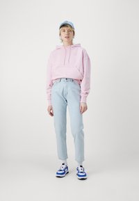 Sudadera rosa clara con bolsillo frontal, gorra azul, jeans de un azul claro y zapatillas deportivas blancas con detalles azules. Diseño sencillo y ajuste casual.