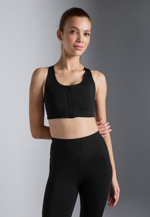 Fekete sportos crop top elöl cipzárral, széles pántokkal és sima textúrával. Magas derekú fekete leggings-szel párosítva.