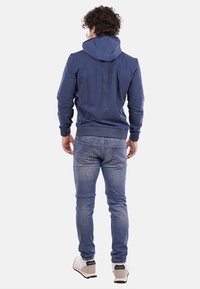 Felpa blu navy con cappuccio regolabile, in tessuto morbido e orlo a coste. Abbinata a jeans in denim blu chiaro e sneakers di colore chiaro.