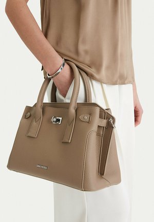 Žena držící béžovou koženou kabelku se stříbrnou přezkou a logem GINO ROSSI, oblečená v béžovém topu, bílých kalhotách a stříbrném náramku.