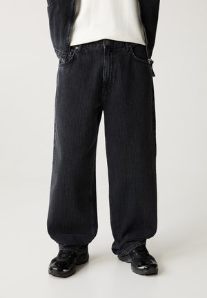 Schwarzweiche Jeans mit weit geschnittenem Bein, vertikaler Rippenstruktur, fünf Taschen und Knopfverschluss. Kombiniert mit schwarzen Sneakern.