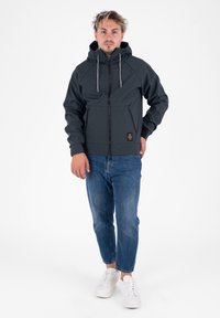 Felpa zip grigia scura con cappuccio, polsini elastici, due tasche frontali e etichetta tessuta. Abbinata a jeans azzurri chiari e sneaker bianche.
