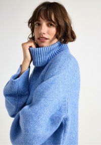 Kvinde med kortbrunt hår iført en lys blå tykk strikket turtleneck sweater, der holder kraven nær sit ansigt, neutral baggrund.