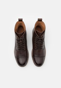 Ett par mörkbruna läder snörade ankstövlar med kapade tår, sedda uppifrån på en vit bakgrund.