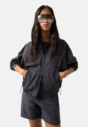 Kvinde med langt mørkt hår iført store reflekterende solbriller, sort letvægts lynlåsjakke og sorte shorts, med hænderne i jakkelommerne.