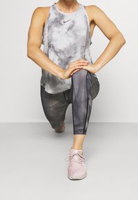 Canotta grigia tie-dye con scollatura rotonda, abbinata a leggings neri semitrasparenti con motivi geometrici e cerniere alla caviglia. Scarpe da ginnastica rosa.