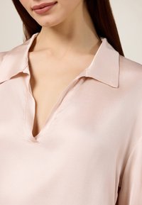 Bluză mătăsoasă, de culoare roz deschis, cu decolteu în V și guler; textură netedă și cusături curate; țesătura se așează ușor în jurul gâtului.