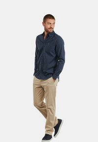 Camicia button-up blu navy con pois bianchi, maniche lunghe e un logo sottile, abbinata a pantaloni khaki e sneakers nere.