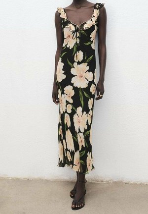 Kobieta ubrana w czarną sukienkę bez rękawów z dużym beżowym kwiatowym wzorem i falbankowymi ramiączkami, zestawioną z czarnymi sandałami na paski.