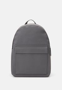Sac à dos gris en tissu lisse, présentant une forme arrondie, une poche avant avec rabat et une poignée sur le dessus. Design minimaliste avec des détails brodés.