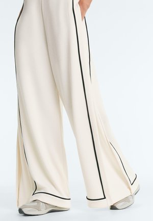 Szerokie kremowe spodnie z czarnym przeszyciem, noszone z beżowo-białymi trampkami, ukazujące swobodny, luźny krój i rękę w kieszeni.