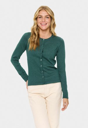 Glimlachende vrouw met lang blond haar, gekleed in een donkergroene knoopcardigan en lichtbeige broek, staand met één hand op de heup.