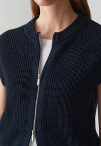 Kvinna som bär en marinblå chunky stickad ärmlös jacka med dragkedja över en vit ribbad topp, som visar halslinje och övre del av torso.