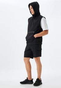 Felpa senza maniche nera con cappuccio e cordino, tasca frontale e pantaloni corti neri. Indossata con scarpe da ginnastica nere. Presenta una vestibilità ampia.
