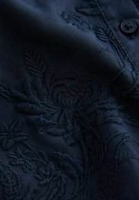 Tissu brodé bleu foncé avec un motif floral, présentant une couture texturée et un détail de bouton. Fini lisse, éclat subtile.