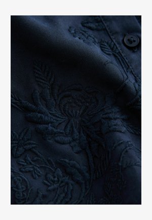 Material brodat de un albastru închis, cu un model floral, având o cusătură texturată și un detaliu cu nasture. Finisaj neted, cu o strălucire subtilă.