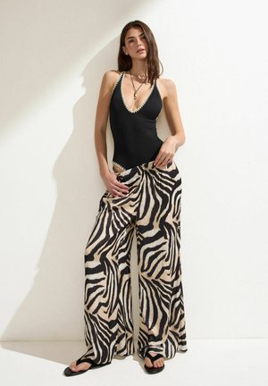 Femme portant un maillot de bain noir à décolleté plongeant avec un bord blanc, un pantalon large à motif zèbre, des sandales noires et un collier pendentif, debout contre un mur blanc.