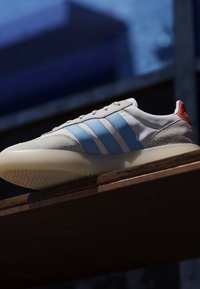 Hvid sneaker med lyseblå striber og rødt hældetalje, der hviler på en træoverflade mod en mørk, sløret baggrund.