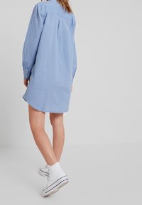 Abito camicia a righe blu e bianche con maniche lunghe, una piega sul retro e un orlo curvo, abbinato a sneaker alte bianche.