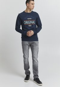 Felpa navy con stampa grafica, colletto tondo, maniche lunghe e polsini a costine. Abbinata a jeans grigi chiari e sneakers nere.