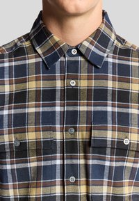 Camicia a quadri nei colori blu navy, giallo e marrone, con colletto button-down. Presenta due taschini sul petto e bottoni grigi. Il tessuto ha una trama texturizzata.