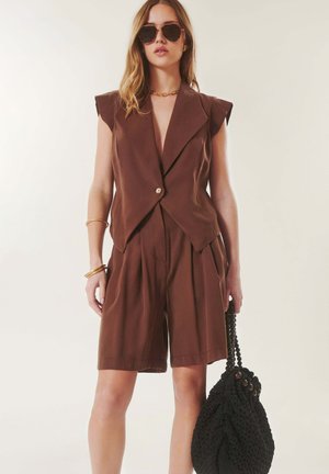 Femme portant un blazer marron sans manches et un short, des lunettes de soleil, des bijoux en or, tenant un sac tressé noir sur un fond clair uni.