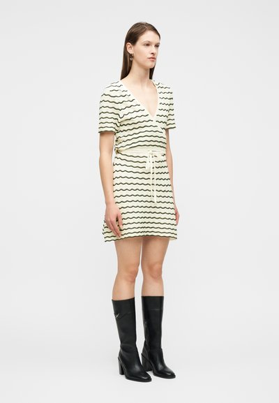 Γυναίκα που φοράει λευκό φόρεμα με μαύρες κυματιστές ρίγες και ζώνη στη μέση, συνδυασμένο με μαύρες ψηλοτάκουνες μπότες μέχρι το γόνατο, στέκεται στο πλάι.