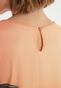Blouse couleur pêche avec une texture lisse, dotée d'une fermeture dos en keyhole maintenue par un bouton. Design minimal, sans motifs visibles.