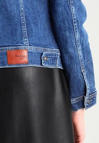 Chaqueta de mezclilla con lavado azul oscuro y un parche de marca de cuero; cuenta con botones a presión y una falda de cuero negra contrastante debajo.