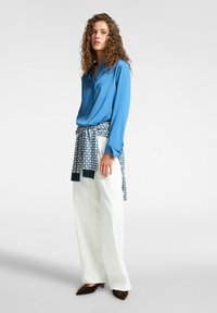 Blusa blu a maniche lunghe in tessuto morbido, abbinata a pantaloni bianchi a gamba larga e un wrap a scacchi. Le scarpe nere a punta completano il look.