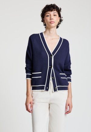 Frau mit kurzen lockigen Haaren, die eine marineblaue Strickjacke mit weißem Besatz und schwarzen Knöpfen trägt, kombiniert mit weißen Hosen, steht vor einem schlichten Hintergrund.